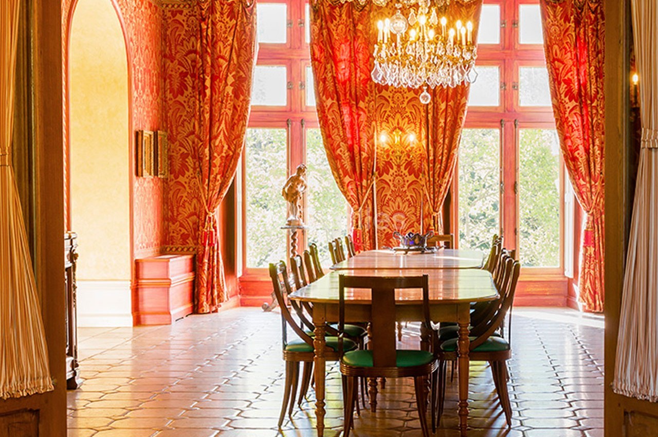 Salle à manger élégante avec une grande table en bois, chaises vertes, lustre en cristal et rideaux rouges ornés.