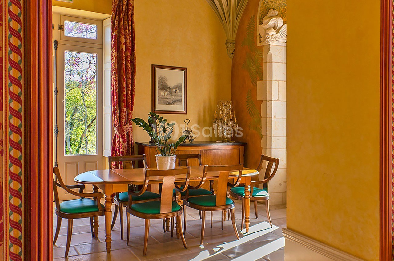 Salle à manger lumineuse avec table en bois, chaises à assise verte, murs jaunes et grandes fenêtres donnant sur un jardin.