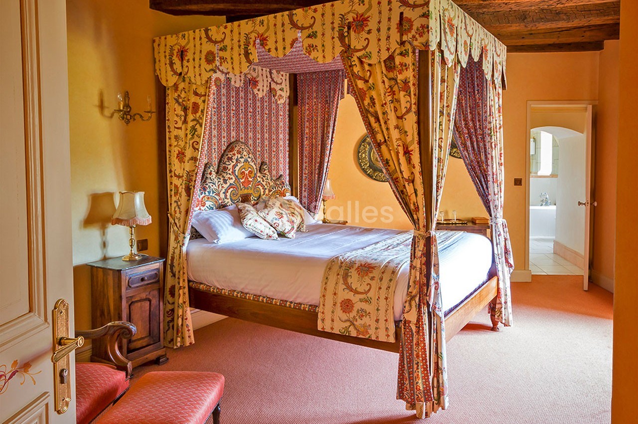 Lit à baldaquin avec tentures colorées dans une chambre chaleureuse aux murs orangés et poutres apparentes.