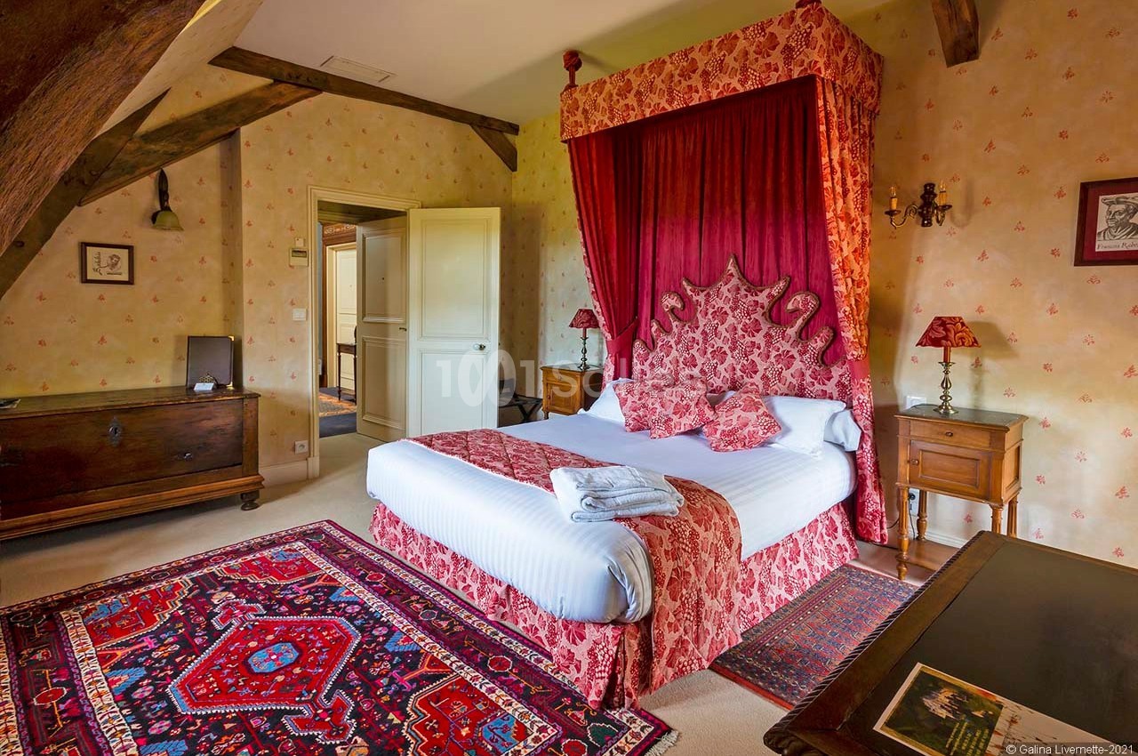 Lit à baldaquin rouge dans une chambre rustique avec poutres apparentes, tapis colorés et meubles en bois.