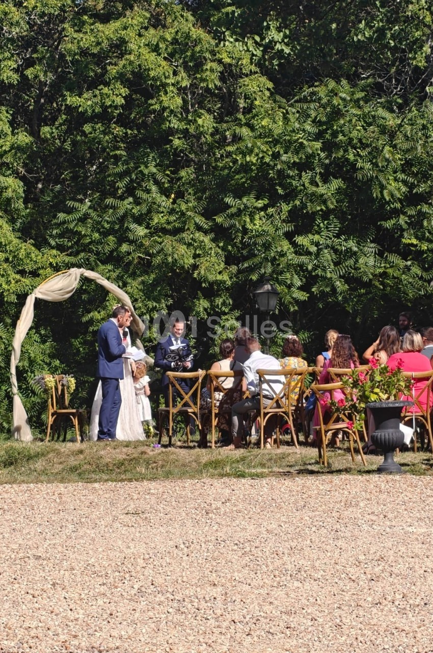 Un couple échange ses vœux sous une arche décorée, entouré d'invités assis en extérieur près d'arbres.