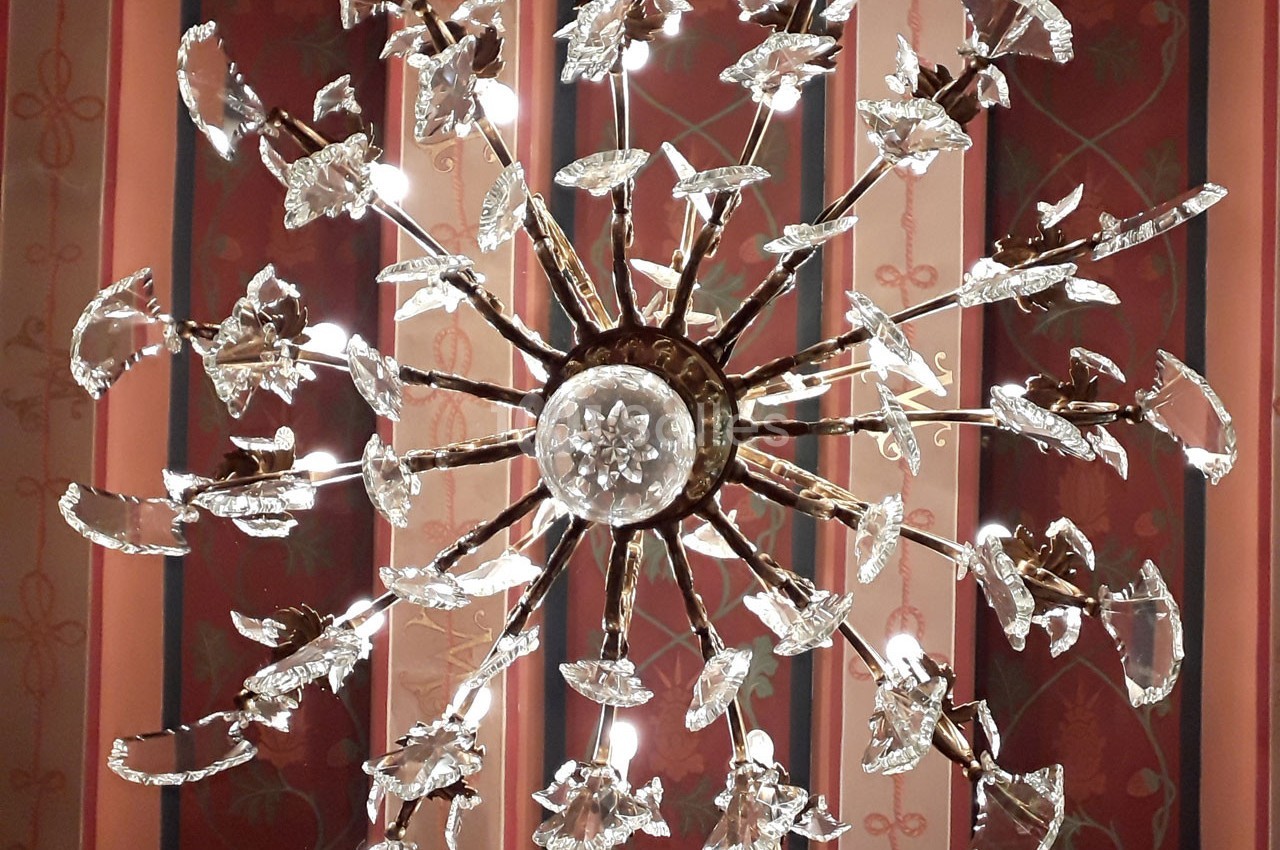 Lustre en métal doré orné de cristaux en forme de fleurs, suspendu devant un mur à motifs rouges et dorés.