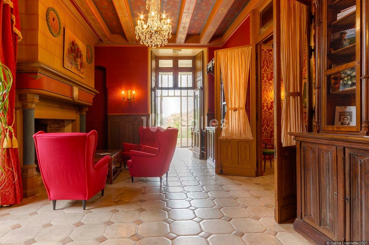Salon élégant avec fauteuils rouges, cheminée, lustre et portes vitrées ouvertes sur un balcon avec vue extérieure.