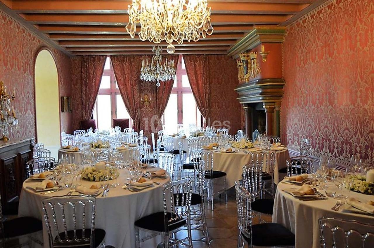 Salle de réception élégante avec tables rondes dressées, chaises transparentes et lustres en cristal.