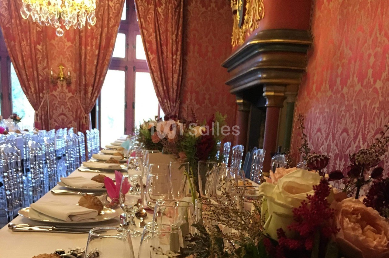 Table de banquet élégamment dressée dans une salle ornée de murs rouges, avec chandeliers et cheminée décorative.