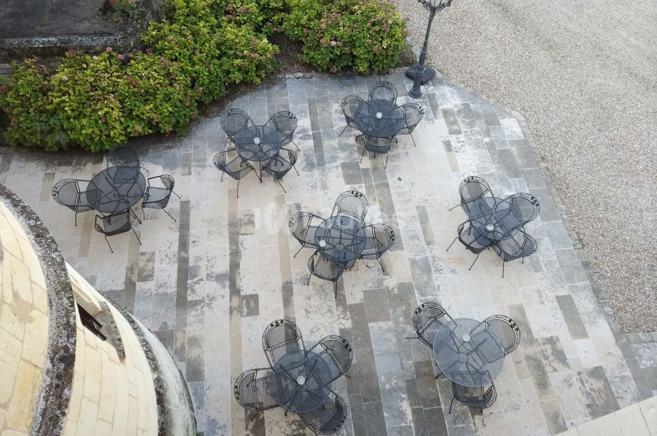 Terrasse pavée avec plusieurs tables et chaises en métal noir, entourée de buissons et d'un lampadaire.