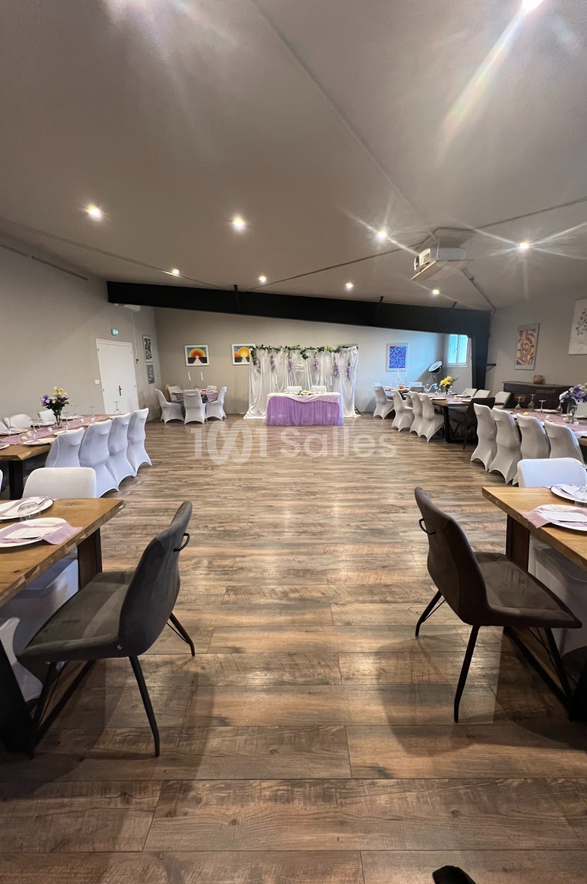 Salle de réception décorée avec des tables dressées, chaises blanches et noires, et une table centrale ornée de fleurs.