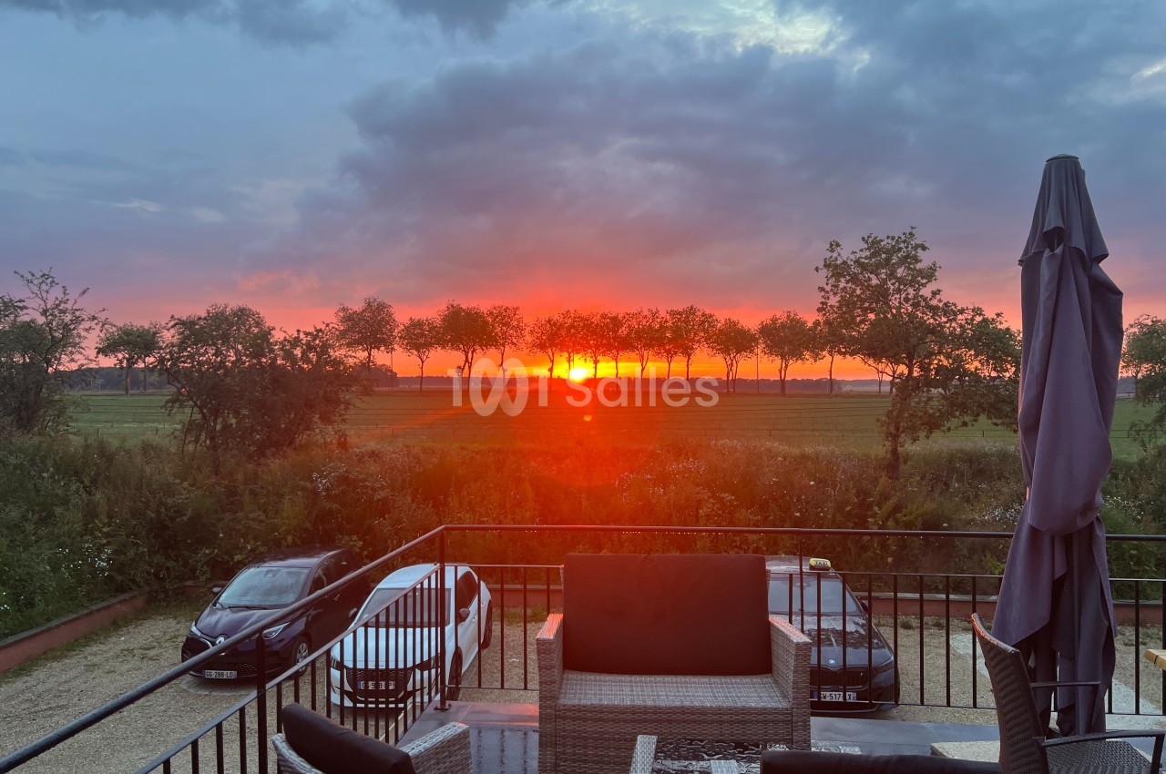 Coucher de soleil rougeoyant derrière une rangée d'arbres, vu depuis une terrasse avec mobilier et voitures garées.