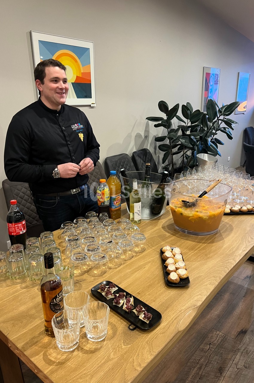 Un homme derrière une table garnie de boissons, verres, amuse-bouches et un grand saladier de cocktail.