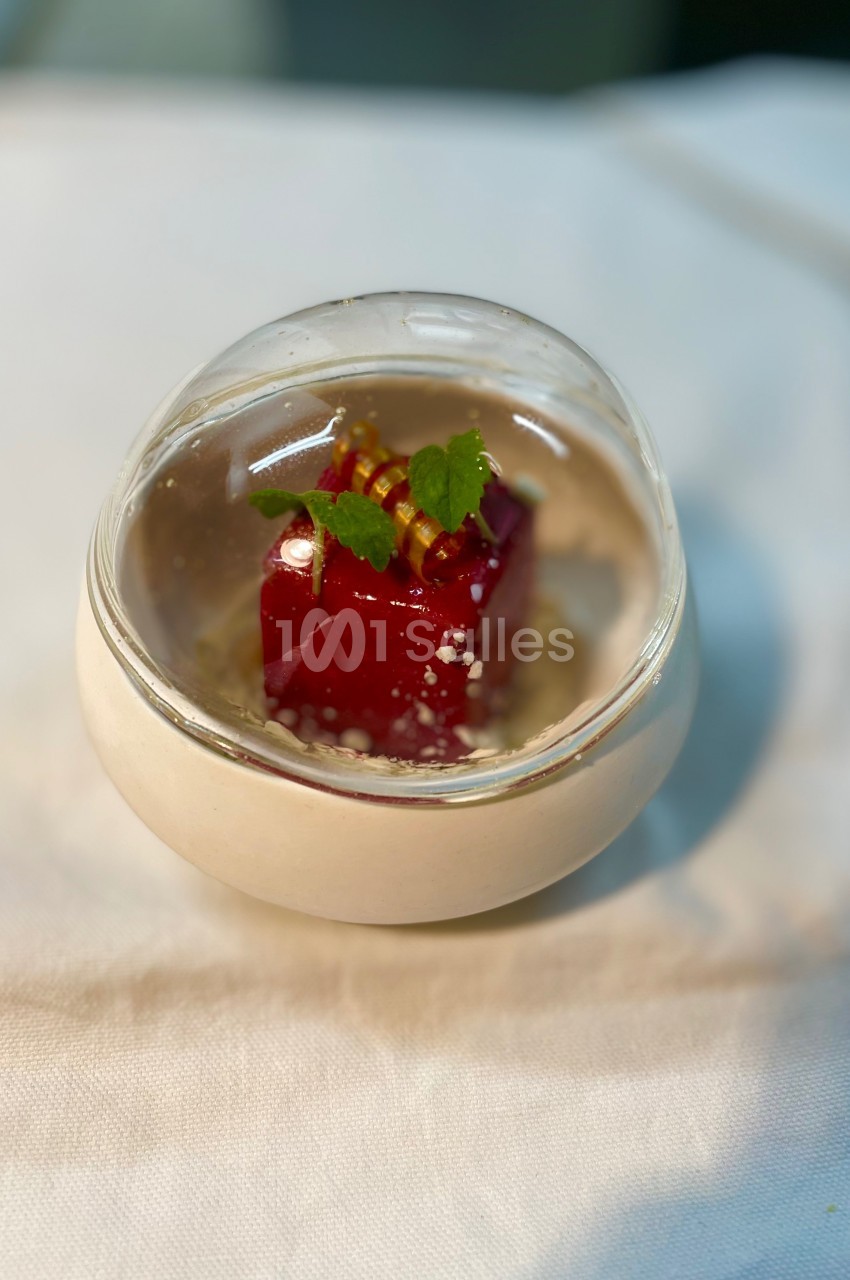 Cube de dessert rouge décoré de feuilles vertes et d'éléments dorés, présenté dans un bol transparent sur une nappe blanche.
