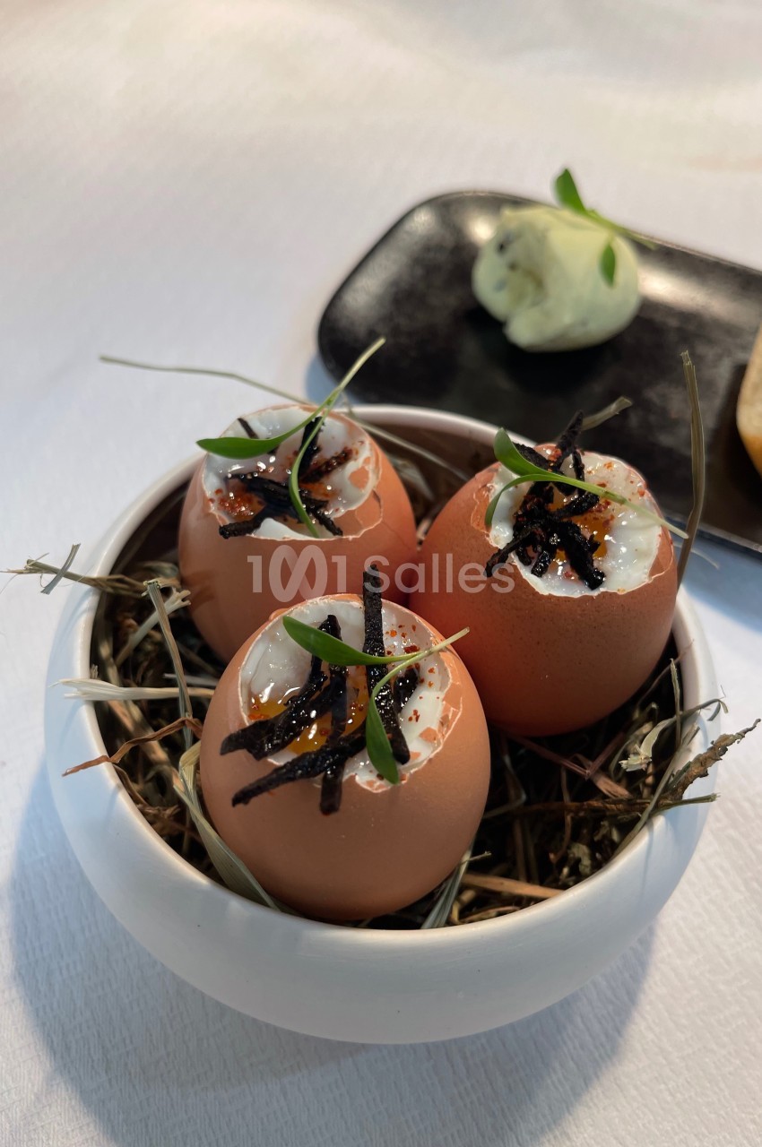 Œufs présentés dans des coquilles, garnis de crème et truffes, disposés sur un lit de foin dans un bol blanc.