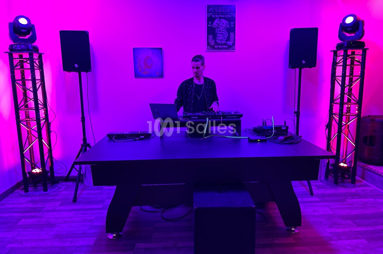 Un DJ mixe sur une table de mixage dans une salle éclairée par des lumières violettes et roses, entouré d'équipements audio.
