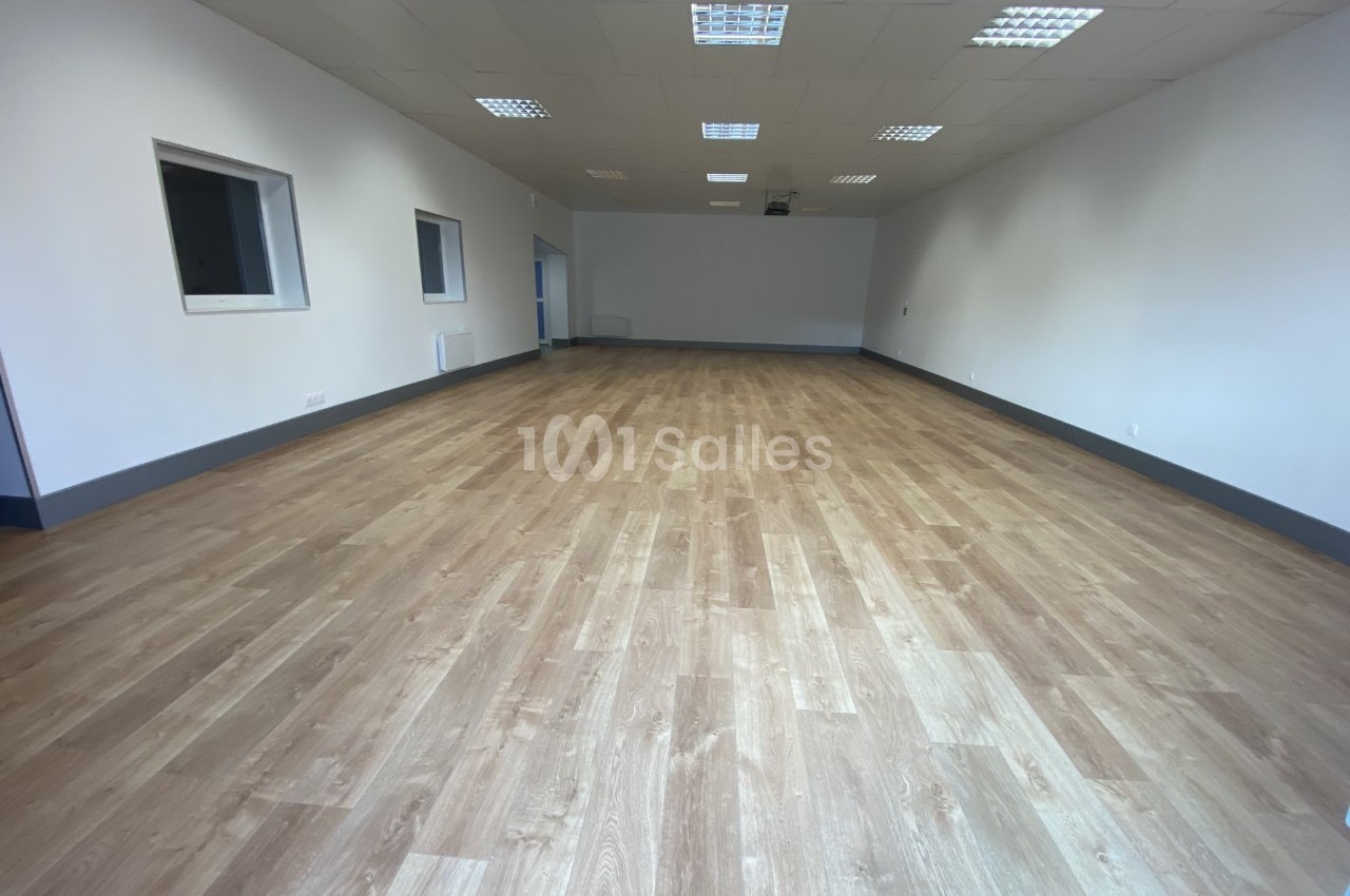 Salle vide avec parquet en bois clair, murs blancs, plafond avec éclairage encastré et fenêtres sur un côté.