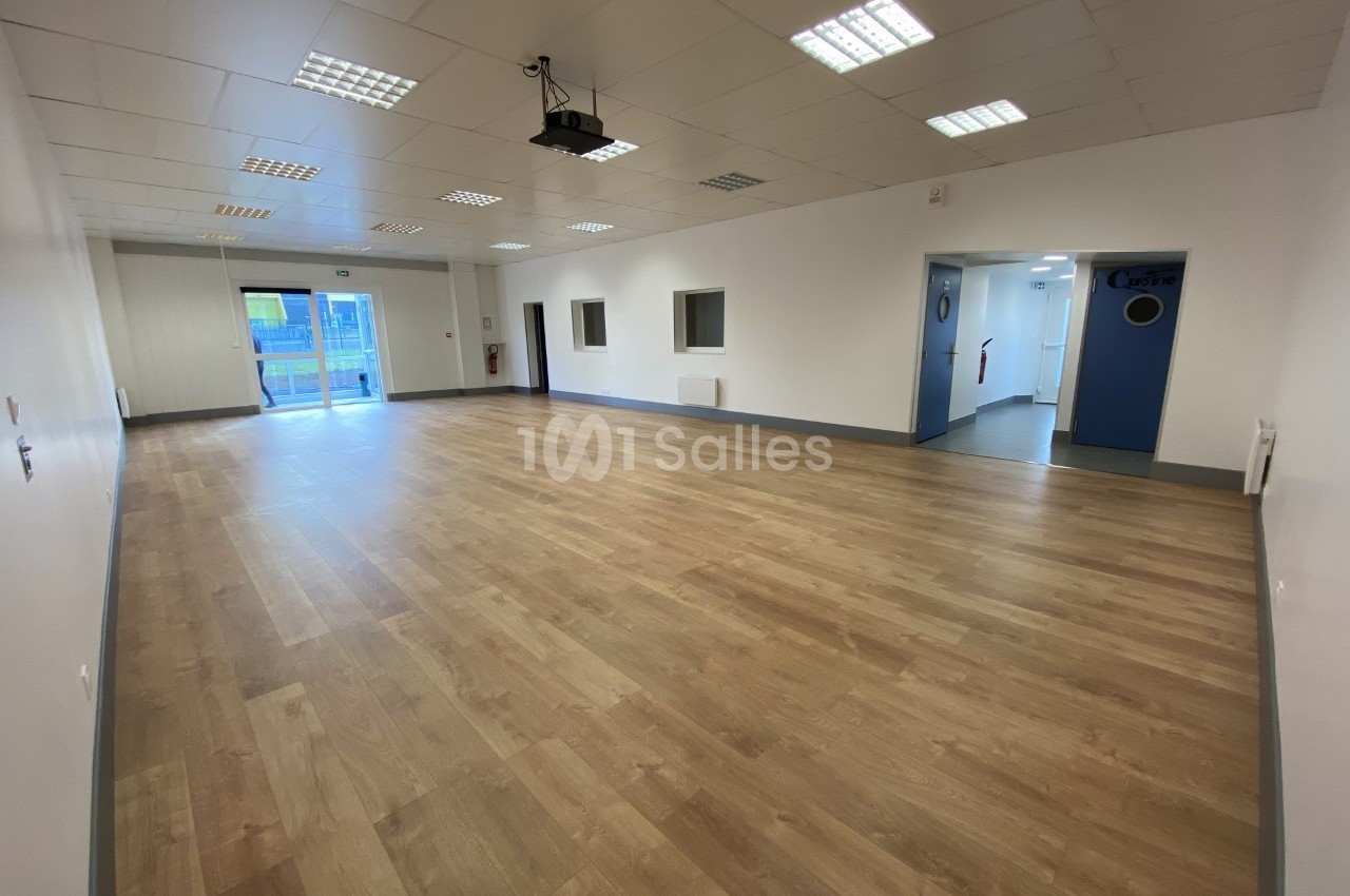 Salle vide avec parquet clair, murs blancs, plafond à dalles et éclairage fluorescent, portes et fenêtres visibles.