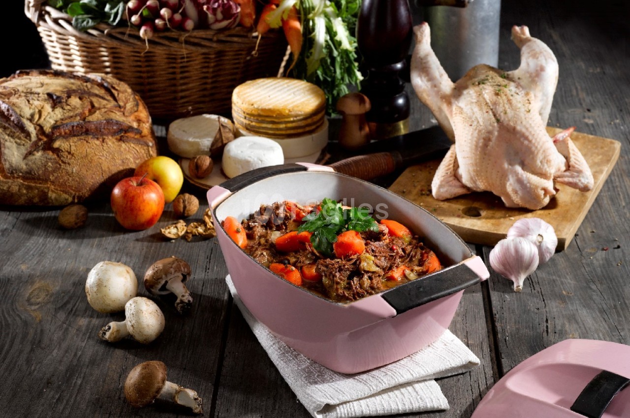 Plat mijoté aux légumes et viande dans une cocotte rose, entouré d'ingrédients frais et d'un poulet cru sur une planche.