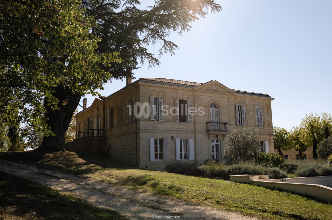 Grande maison en pierre de style classique entourée d'arbres et d'un jardin sous une lumière ensoleillée.