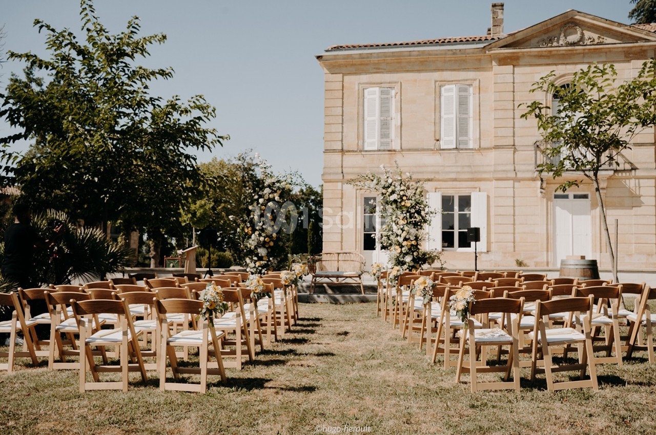 Chaises en bois disposées en rangées devant une maison en pierre, décorées pour une cérémonie en extérieur.