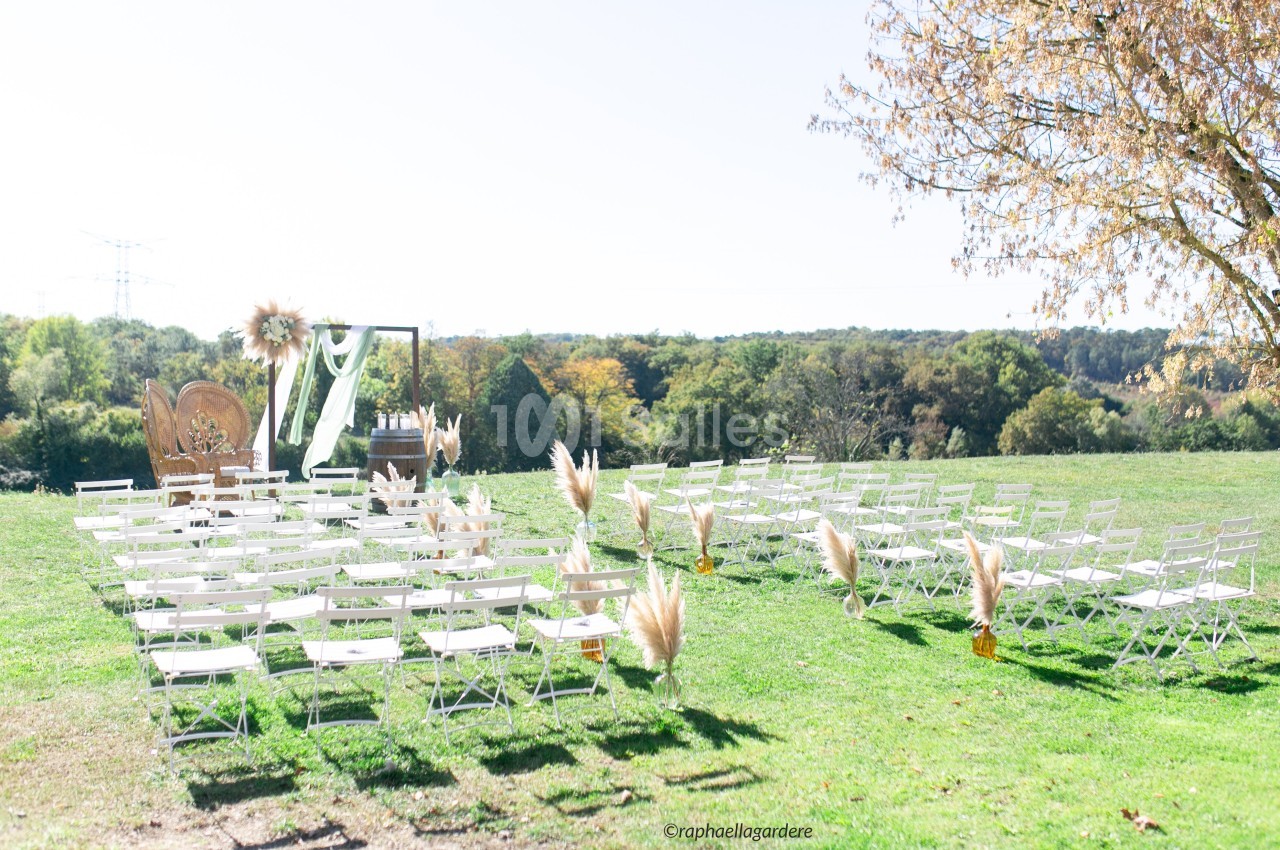Chaises blanches disposées en rangées sur une pelouse pour une cérémonie en plein air, avec arche décorée et paysage boisé.
