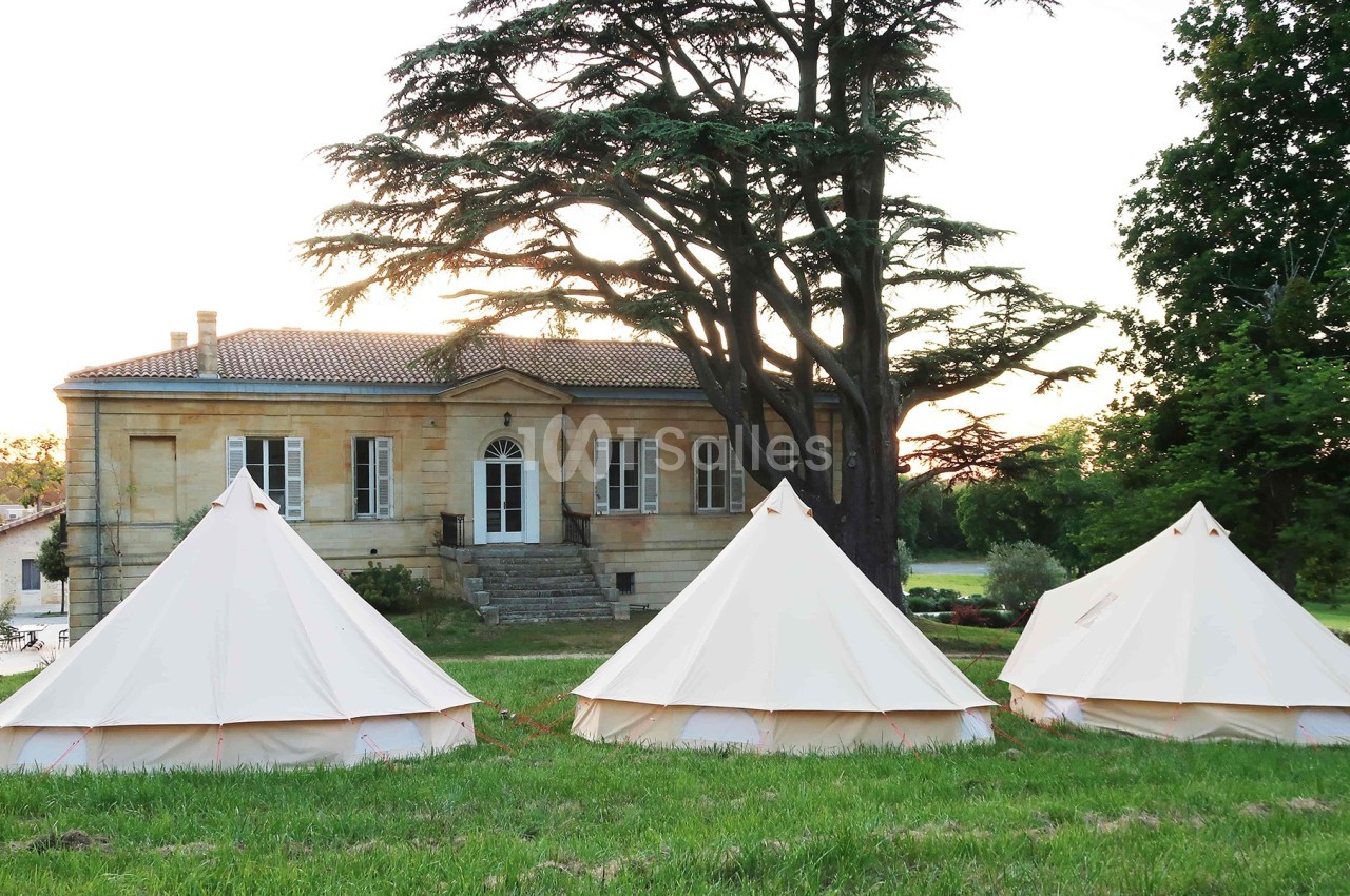 Trois tentes blanches installées sur une pelouse devant une maison ancienne entourée d'arbres au coucher du soleil.