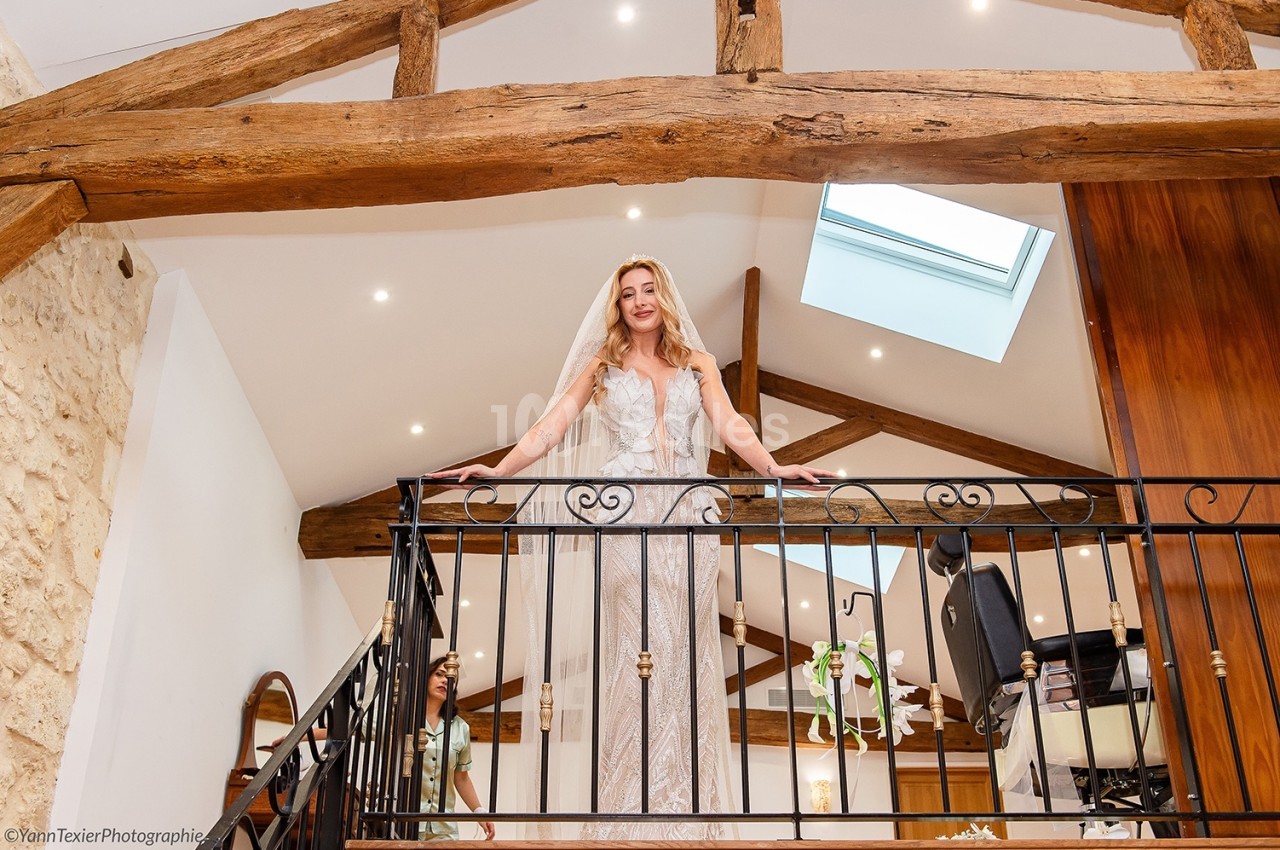 Femme en robe de mariée souriant depuis une mezzanine avec poutres apparentes et lumière naturelle.