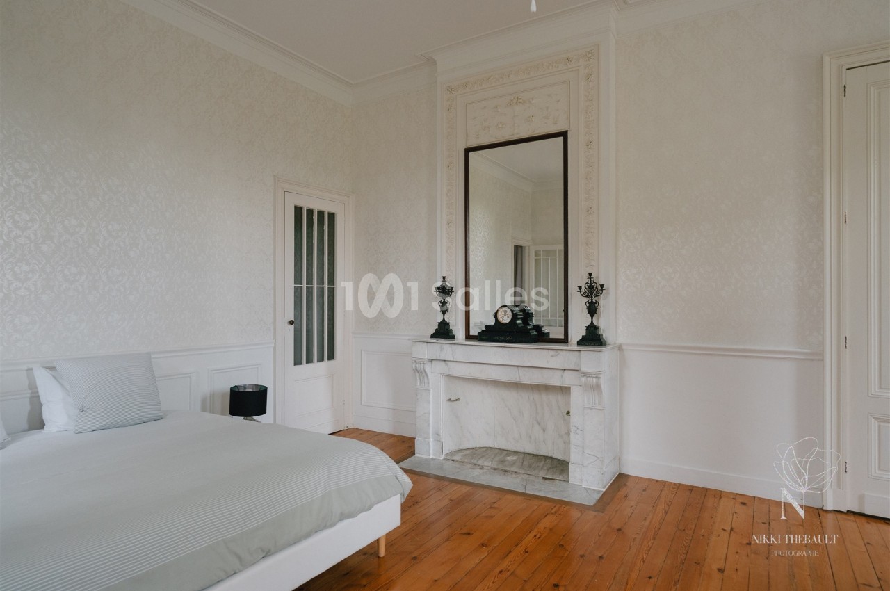 Chambre lumineuse avec lit, cheminée en marbre blanc, grand miroir et parquet en bois clair.