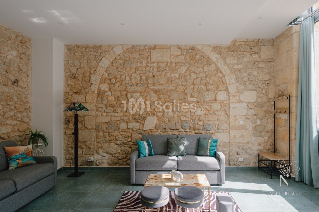 Salon lumineux avec murs en pierre apparente, canapé gris, table basse et grandes fenêtres laissant entrer la lumière.