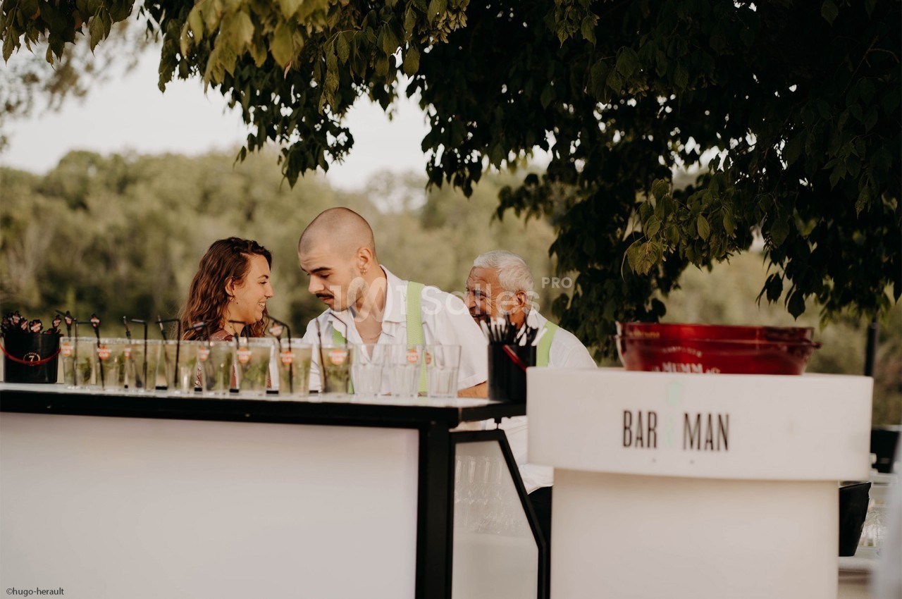 Trois personnes derrière un bar extérieur, entourées de verres et d'arbres en arrière-plan.