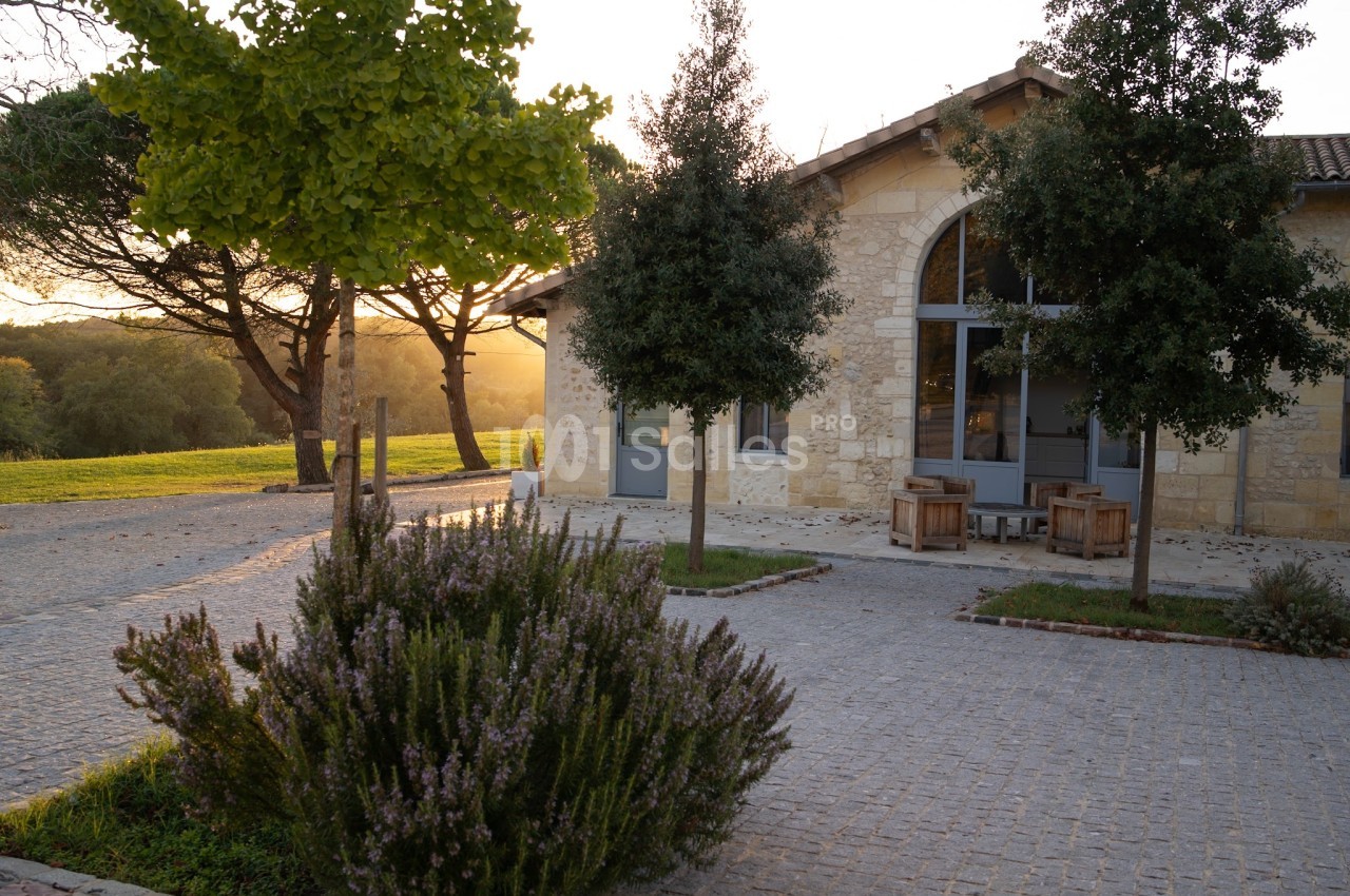 Façade d'un bâtiment en pierre avec grandes fenêtres, entouré d'arbres et d'un espace pavé au coucher du soleil.