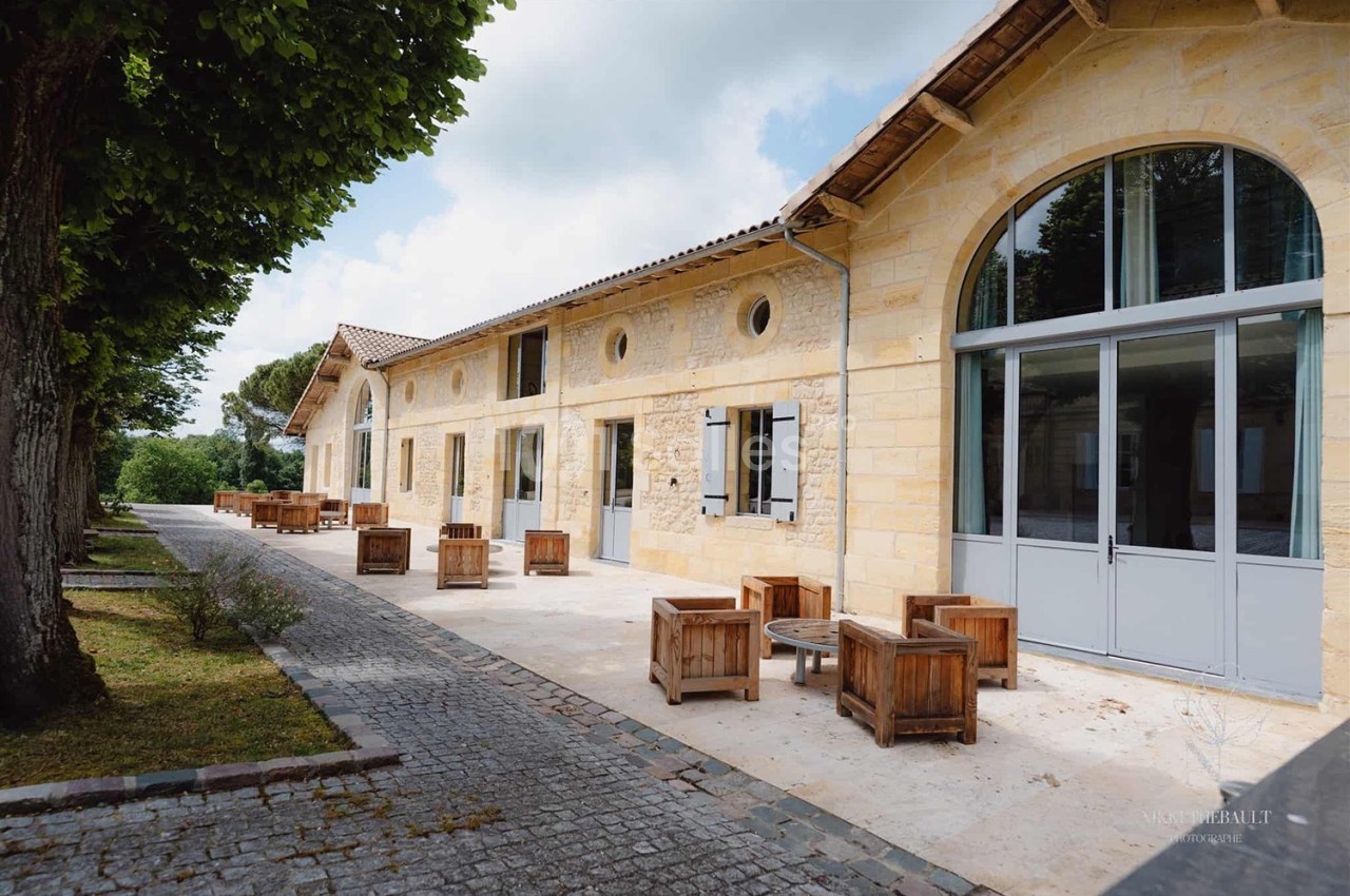 Façade en pierre d'un bâtiment avec terrasse aménagée de fauteuils en bois, entourée d'arbres et d'un chemin pavé.