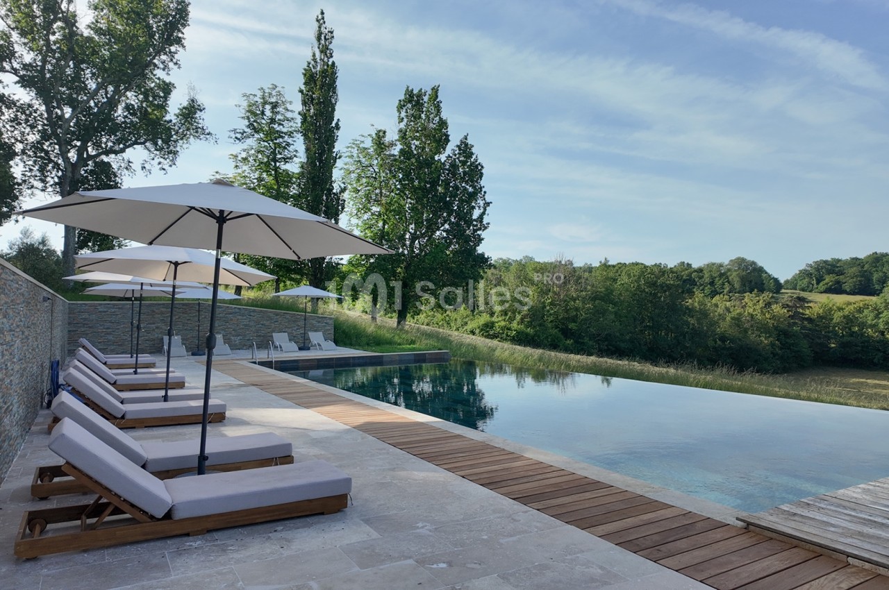 Piscine à débordement entourée de chaises longues et parasols, avec vue sur un paysage verdoyant.
