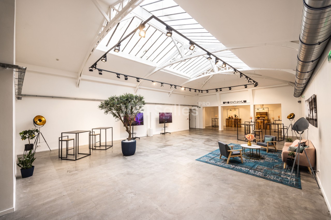 Salle lumineuse avec verrière, mobilier moderne, canapé, tapis bleu et plantes, aménagée pour un événement ou une exposition.