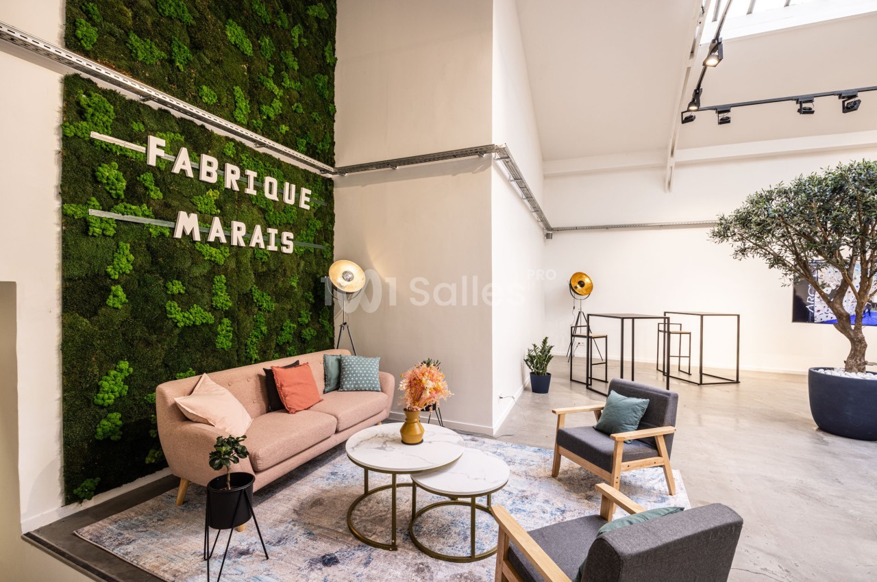 Espace lumineux avec canapé, fauteuils, tables basses et mur végétal portant l'inscription ’Fabrique Marais’.