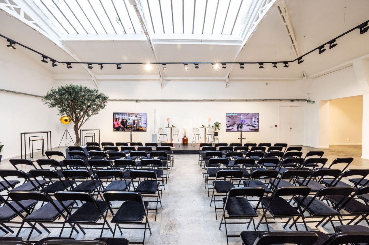 Salle lumineuse avec des rangées de chaises noires, un podium central et deux écrans sous une verrière.
