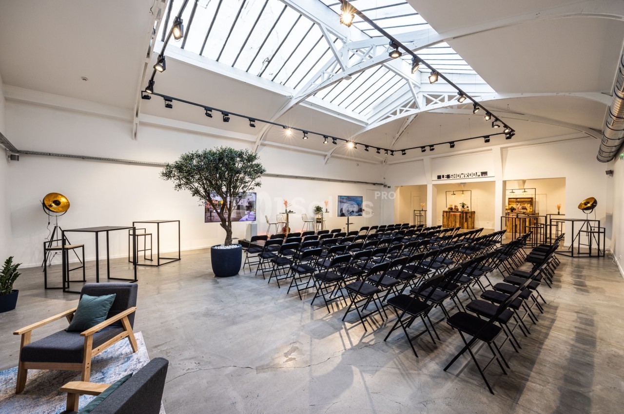 Salle lumineuse avec verrière, rangées de chaises noires, un olivier en pot et un espace aménagé en fond.