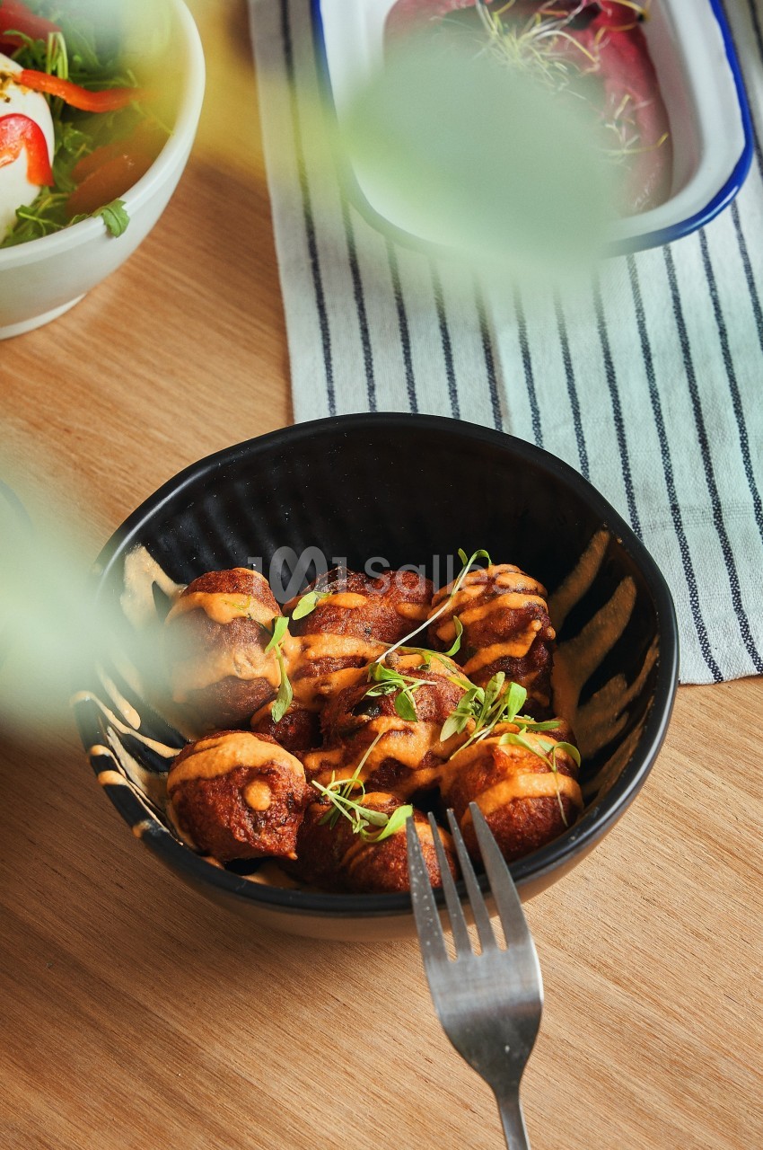 Bol de bouchées dorées garnies de sauce et de micro-pousses, posé sur une table en bois avec des plats en arrière-plan.