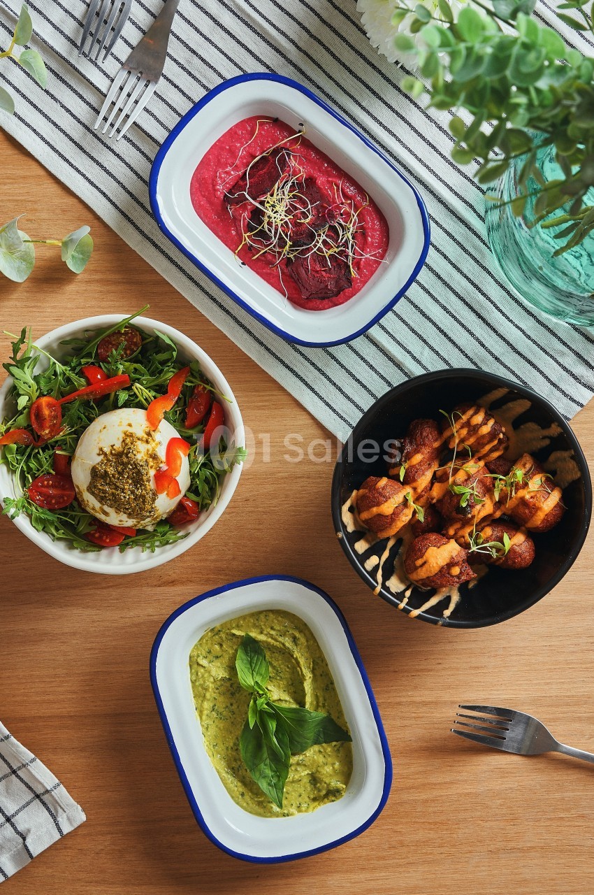 Quatre plats variés sur une table en bois : salade, houmous, brochettes et tartinade rouge, avec des couverts et une nappe.
