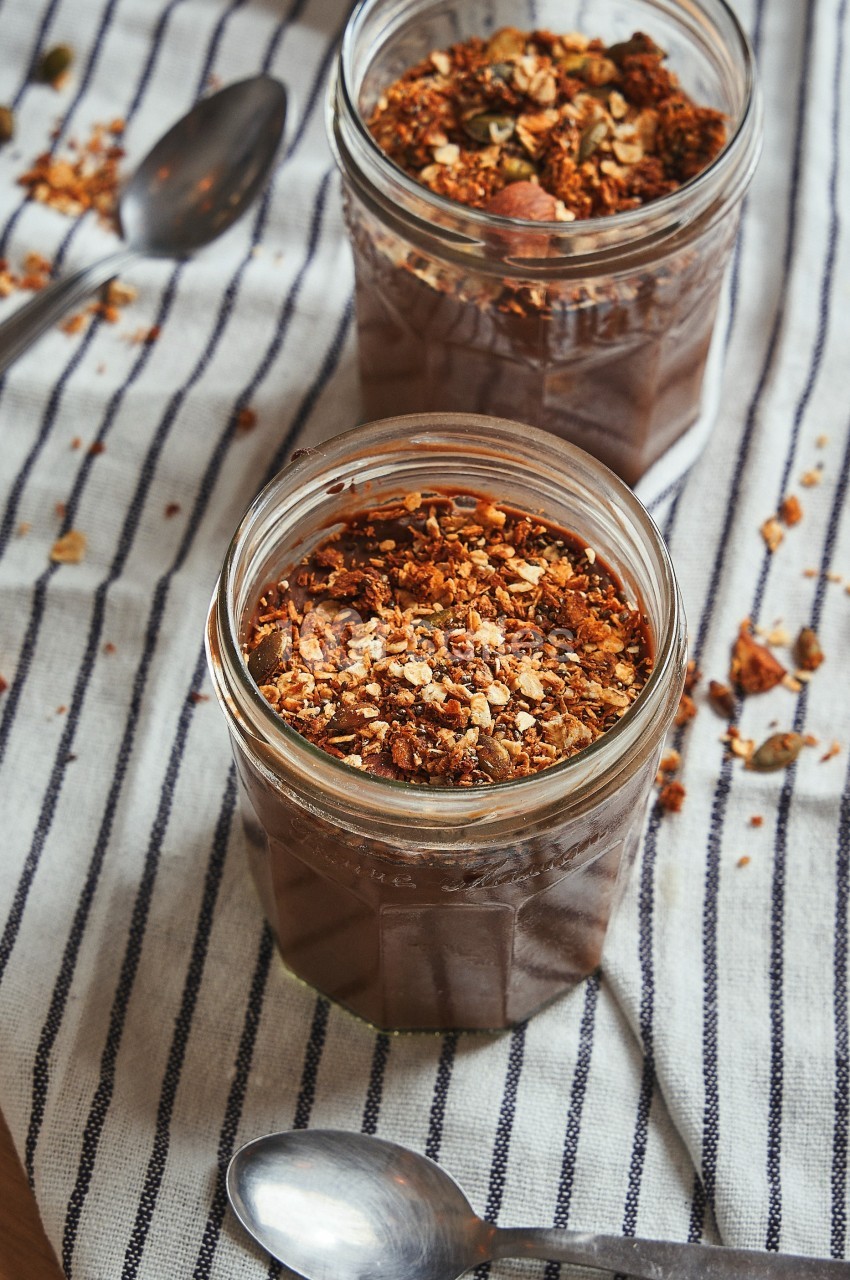 Deux pots de dessert au chocolat garnis de granola, posés sur un torchon rayé avec des cuillères à côté.