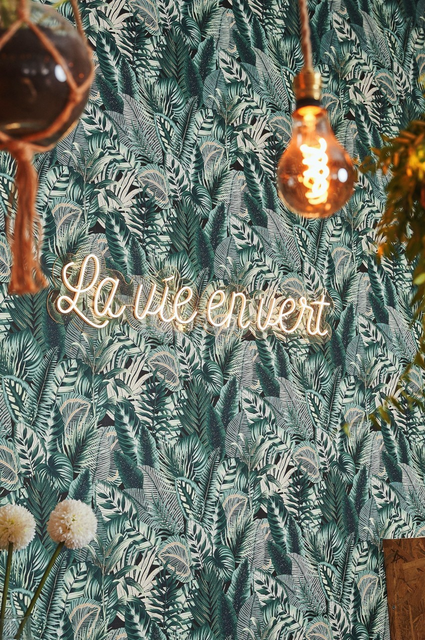 Mur décoré d'un papier peint à motif végétal, éclairé par une enseigne néon ’La vie en vert’ et des suspensions lumineuses.
