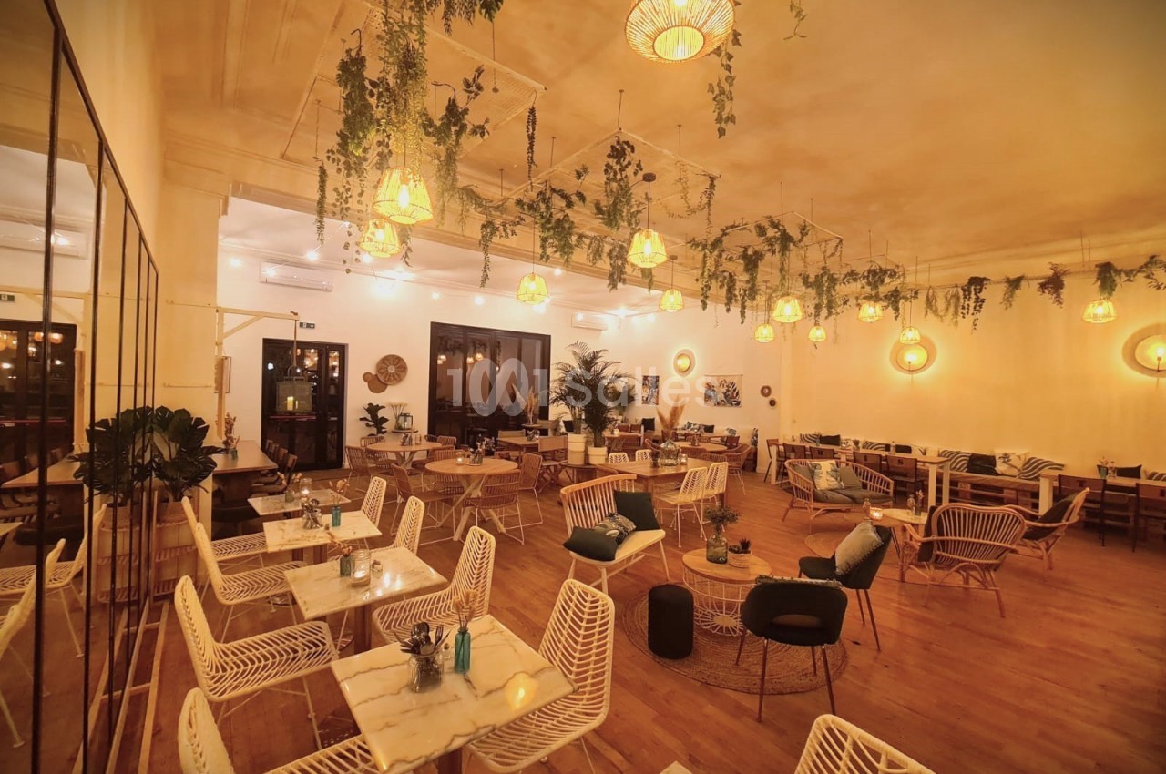 Salle de restaurant chaleureuse avec mobilier en rotin, tables en marbre, éclairage tamisé et plantes suspendues.