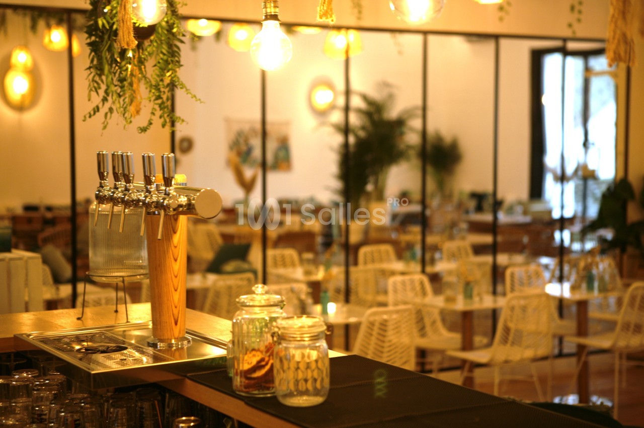 Comptoir de bar avec tireuses à bière, pots en verre et vue sur une salle lumineuse avec des tables et des plantes.