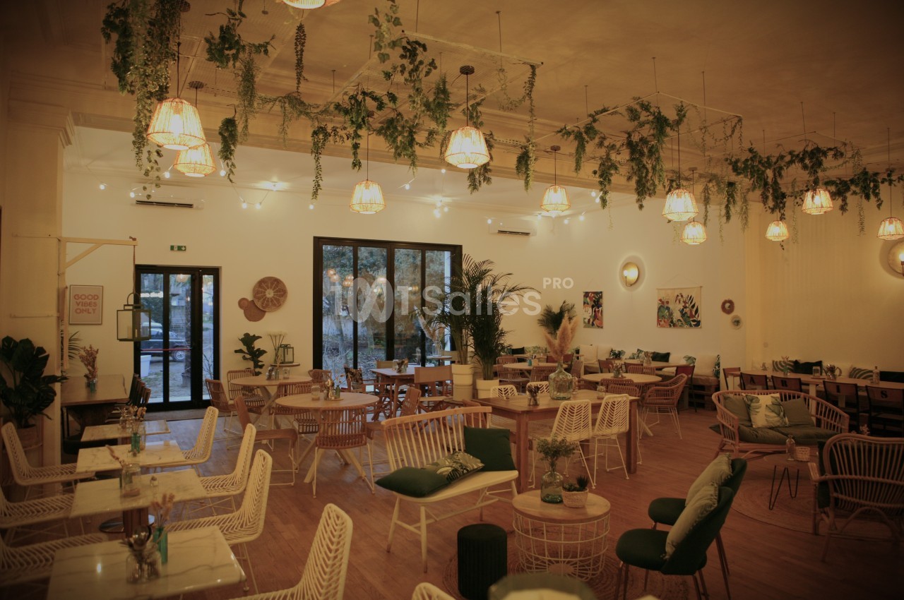 Salle de restaurant chaleureuse avec mobilier en rotin, plantes suspendues et éclairage tamisé.