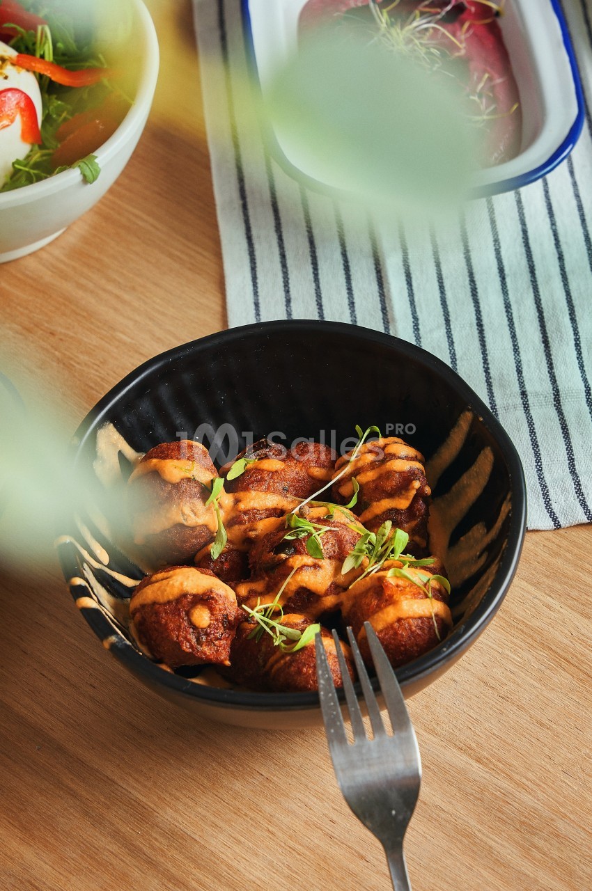 Bol de bouchées dorées garnies de sauce et de micro-pousses, posé sur une table en bois avec des plats en arrière-plan.