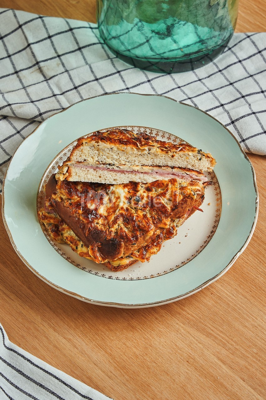 Part de croque-monsieur gratiné sur une assiette blanche bordée de motifs, posée sur une table en bois avec une nappe à…