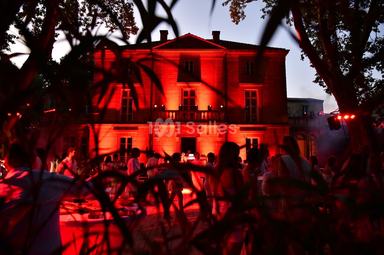 Façade d'un bâtiment éclairé en rouge au crépuscule, entouré d'arbres et d'une foule en extérieur.