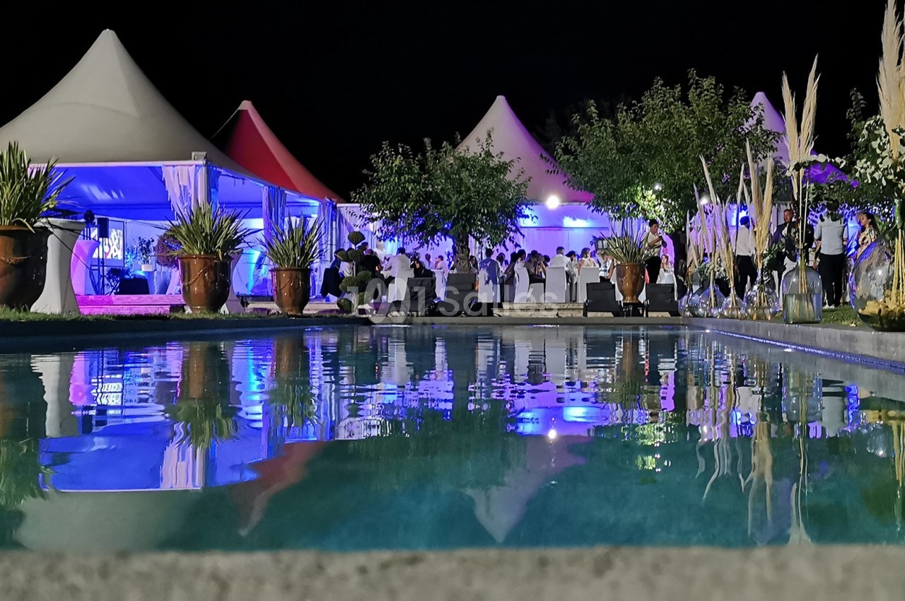 Vue nocturne d'une réception en plein air avec des tentes éclairées, des invités et une piscine au premier plan.
