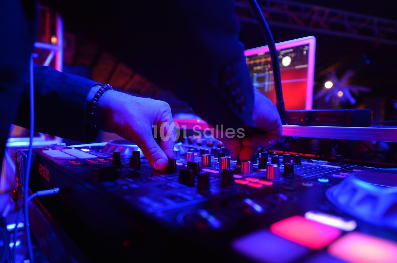 Un DJ ajuste les commandes d'une table de mixage éclairée par des lumières colorées dans un environnement sombre.
