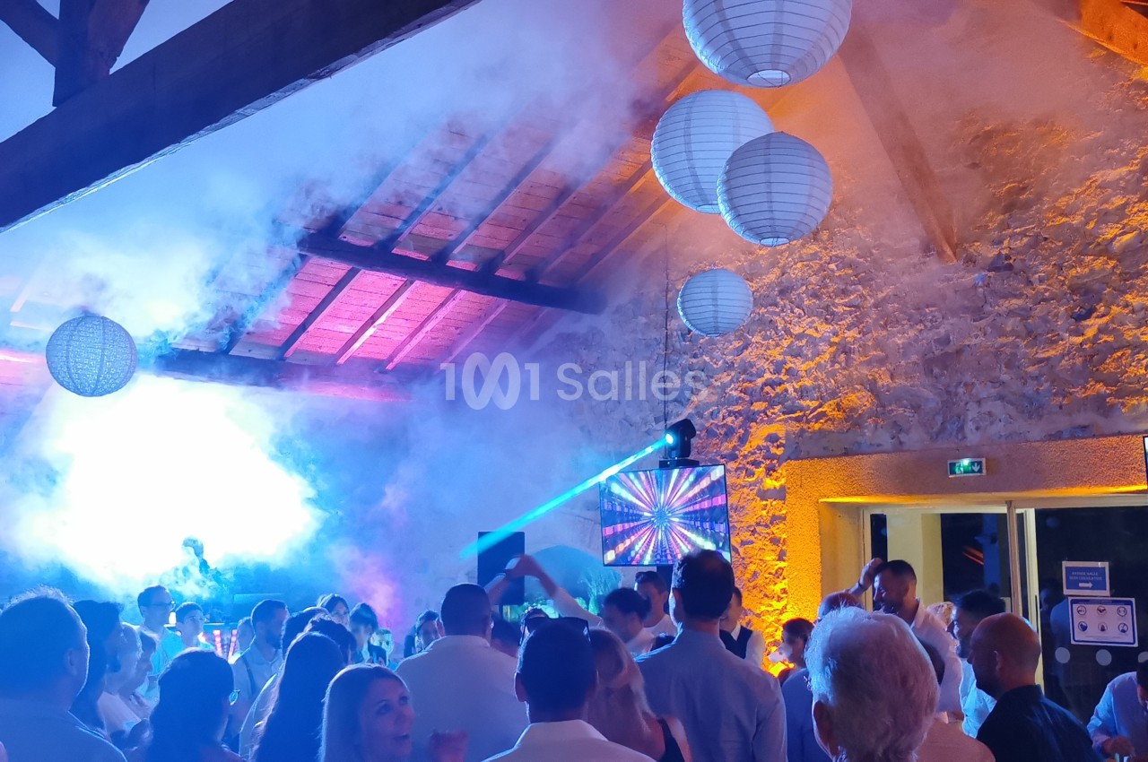 Salle en pierre illuminée avec des lanternes suspendues, une foule dansant sous des jeux de lumière et de la fumée.
