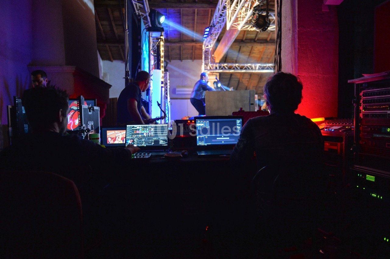 Techniciens supervisant des équipements audiovisuels en coulisses lors d'un événement avec scène éclairée en arrière-plan.