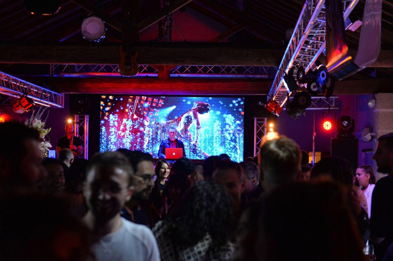 Un DJ mixe sur scène devant un écran lumineux, entouré d'une foule dans une ambiance festive et colorée.
