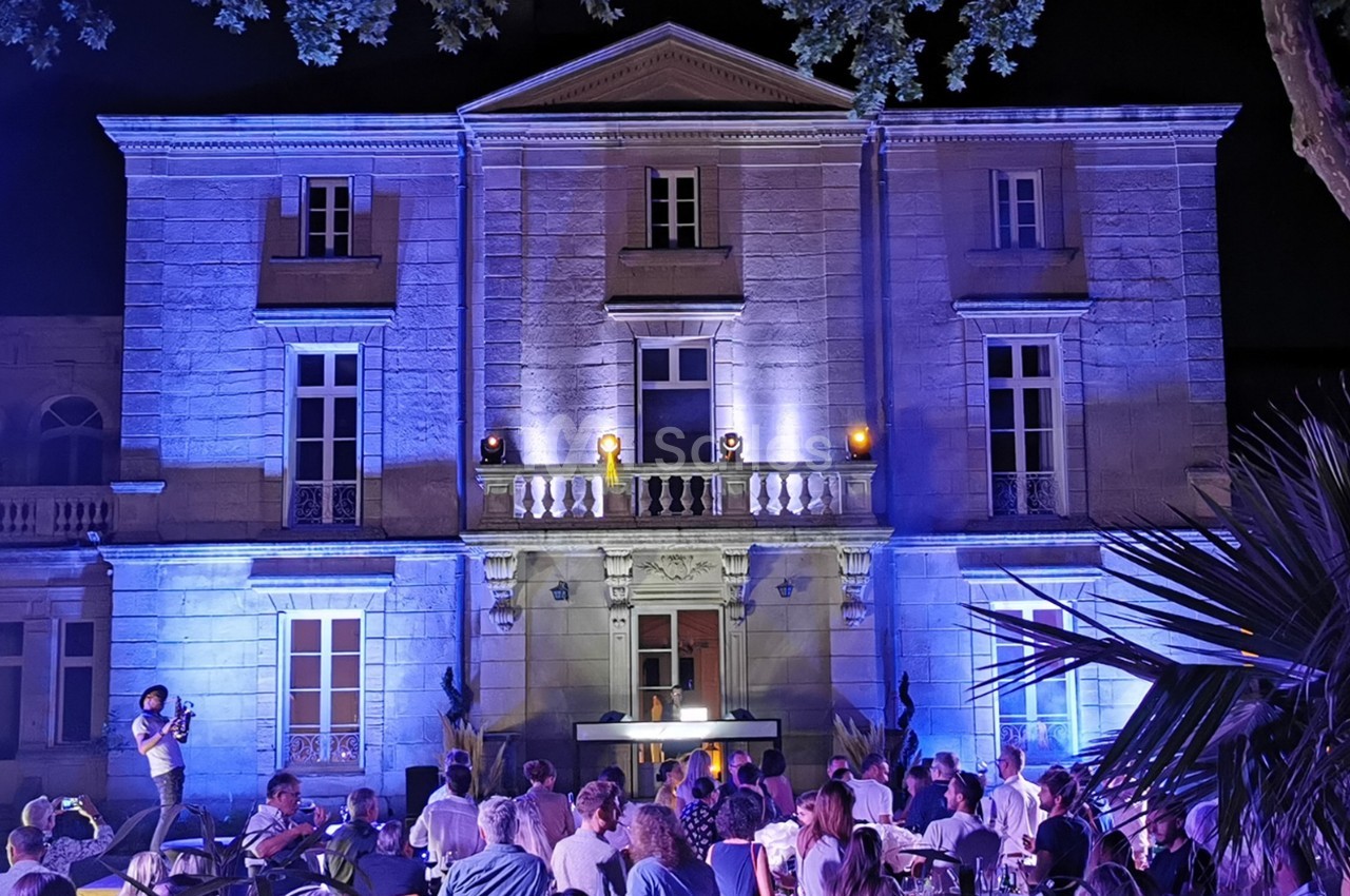 Façade d'un bâtiment éclairé en bleu la nuit, avec des personnes assises devant lors d'un événement en extérieur.